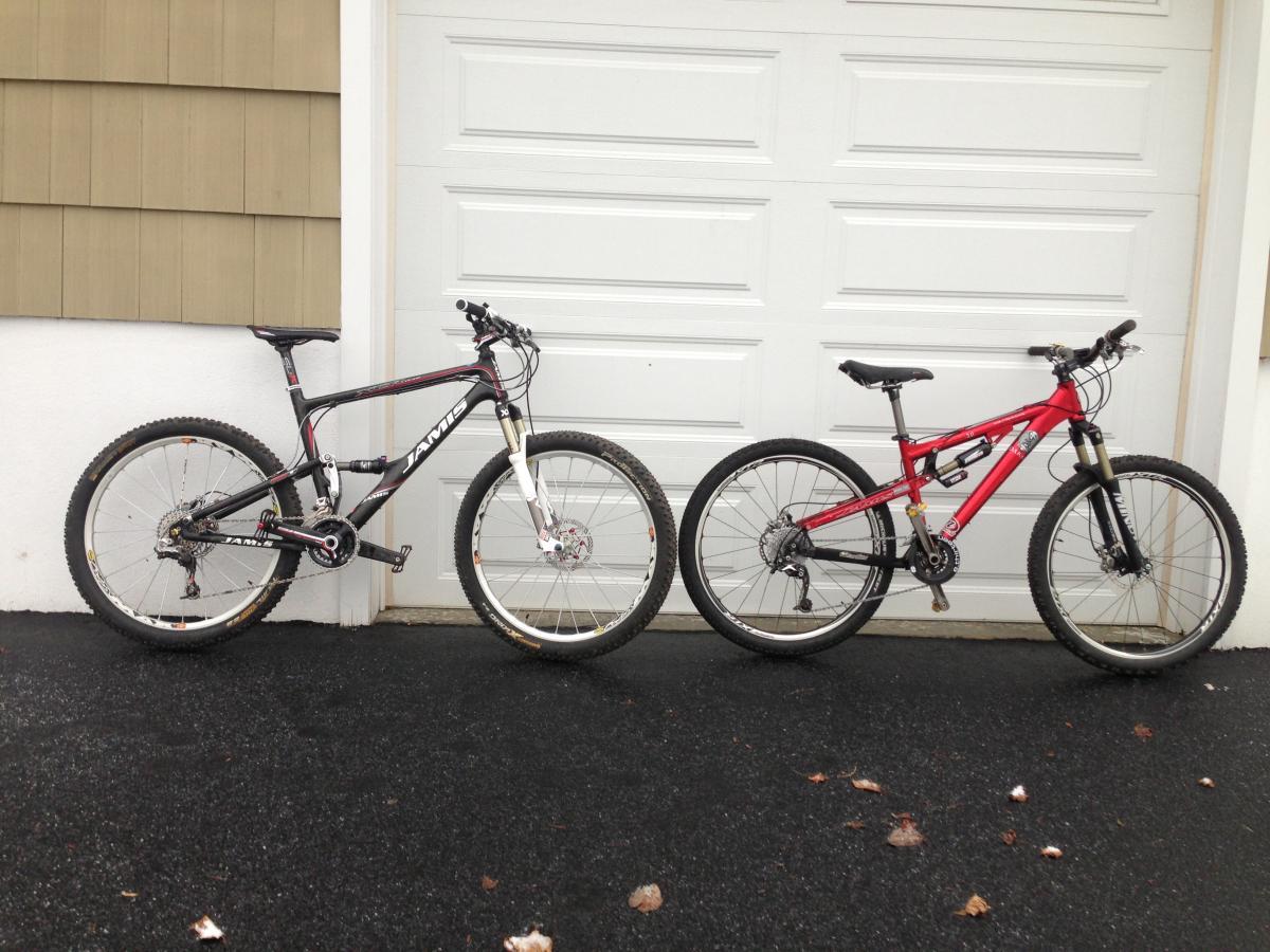 Jamis Dakar XCR Comp: Two mountain bikes are positioned side by side on a dark asphalt surface in front of a white garage door. The left bike features a black frame with red and white accents, while the right bike has a vibrant red frame. Both bikes have rugged tires and are equipped with front suspension forks, indicating their suitability for off-road cycling.