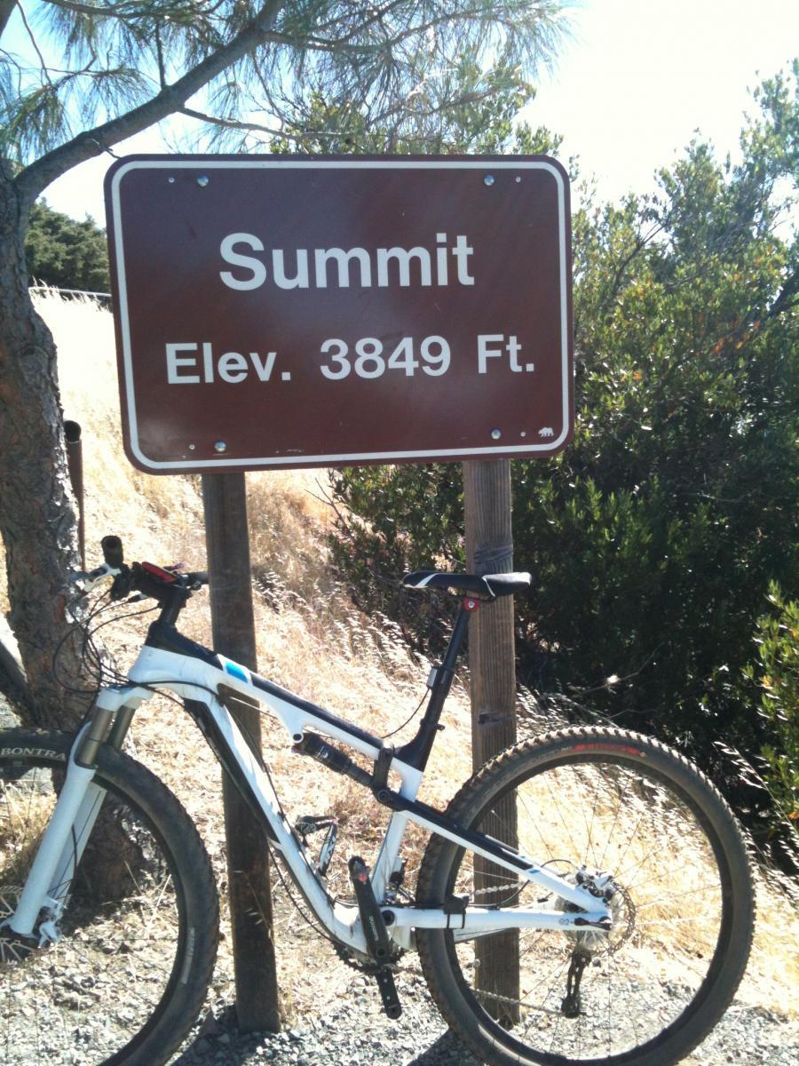 Trek Superfly Full Suspension: A mountain bike parked beside a brown sign that states "Summit" and "Elev. 3849 Ft." The background features dry grass and green shrubbery under a clear blue sky.