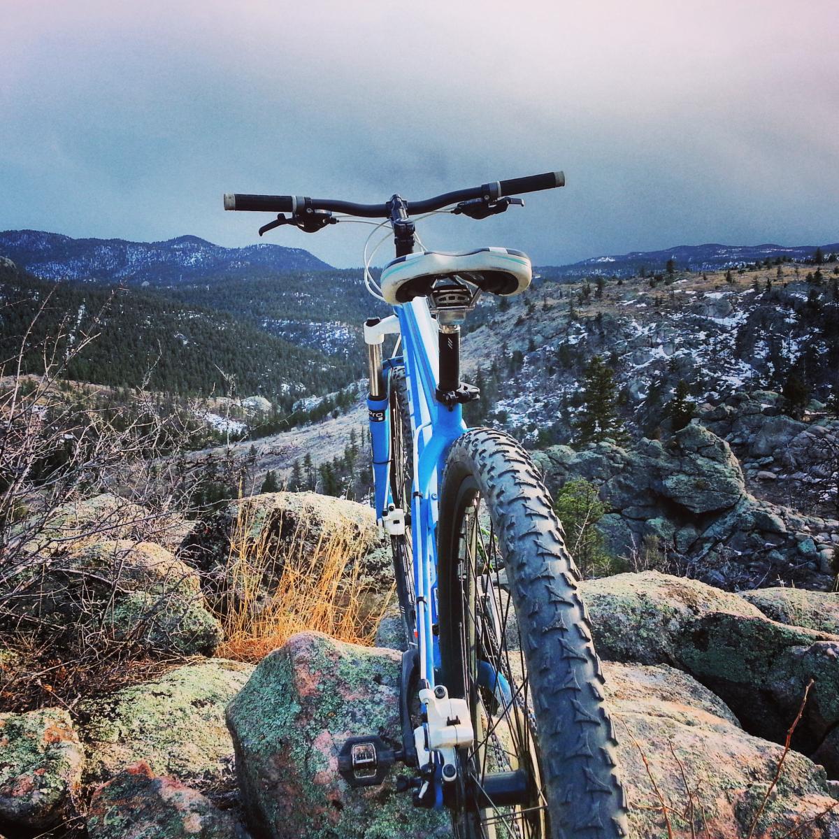 Trek Wahoo: A blue mountain bike resting on rocky terrain, with a scenic view of mountains and valleys in the background. The sky appears overcast, suggesting a dramatic atmosphere, while the landscape is dotted with trees and patches of snow.