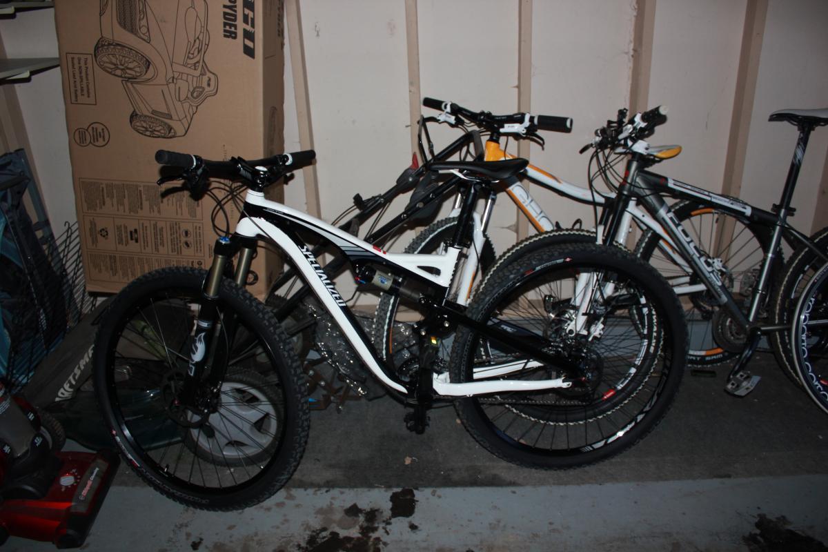 Specialized Camber 29: A black and white mountain bike is parked in a garage next to two other bicycles, with a cardboard box and various items in the background. The garage has concrete flooring and wooden paneling along the walls.