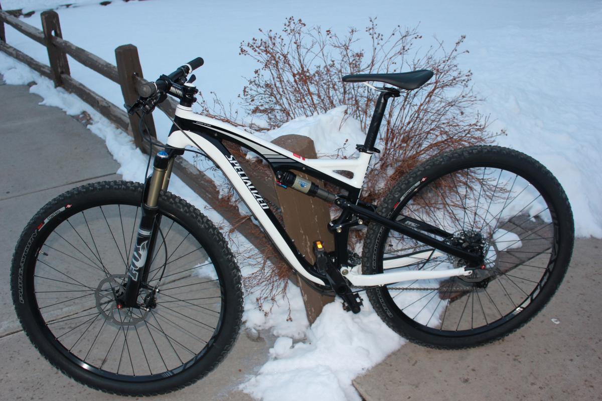 Specialized Camber 29: A black and white mountain bike parked beside a wooden post, with snow and a concrete pathway in the background.