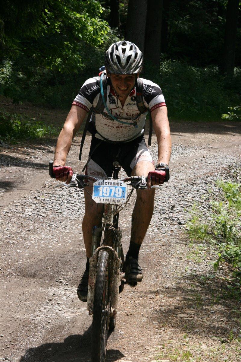 A muddy cyclist on a mountain bike, smiling as they ride along a gravel trail surrounded by trees. The cyclist is wearing a helmet and a dirt-stained jersey with the number 1979.