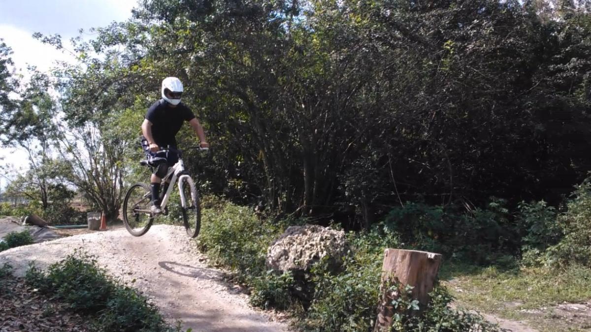 A person in a white helmet is jumping a dirt ramp on a mountain bike, surrounded by lush greenery and trees. The rider is wearing a black shirt and knee pads, with a trail and large rocks visible in the background. Amelia Earhart Park mountain bike trail.