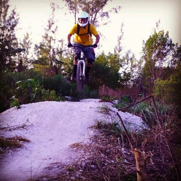 A mountain biker in a yellow shirt and helmet is mid-air, jumping off a dirt ramp in a wooded area. Surrounding trees and vegetation provide a natural backdrop. The biker is focused and wearing protective gear, showcasing an adventurous spirit. Amelia Earhart Park mountain bike trail.