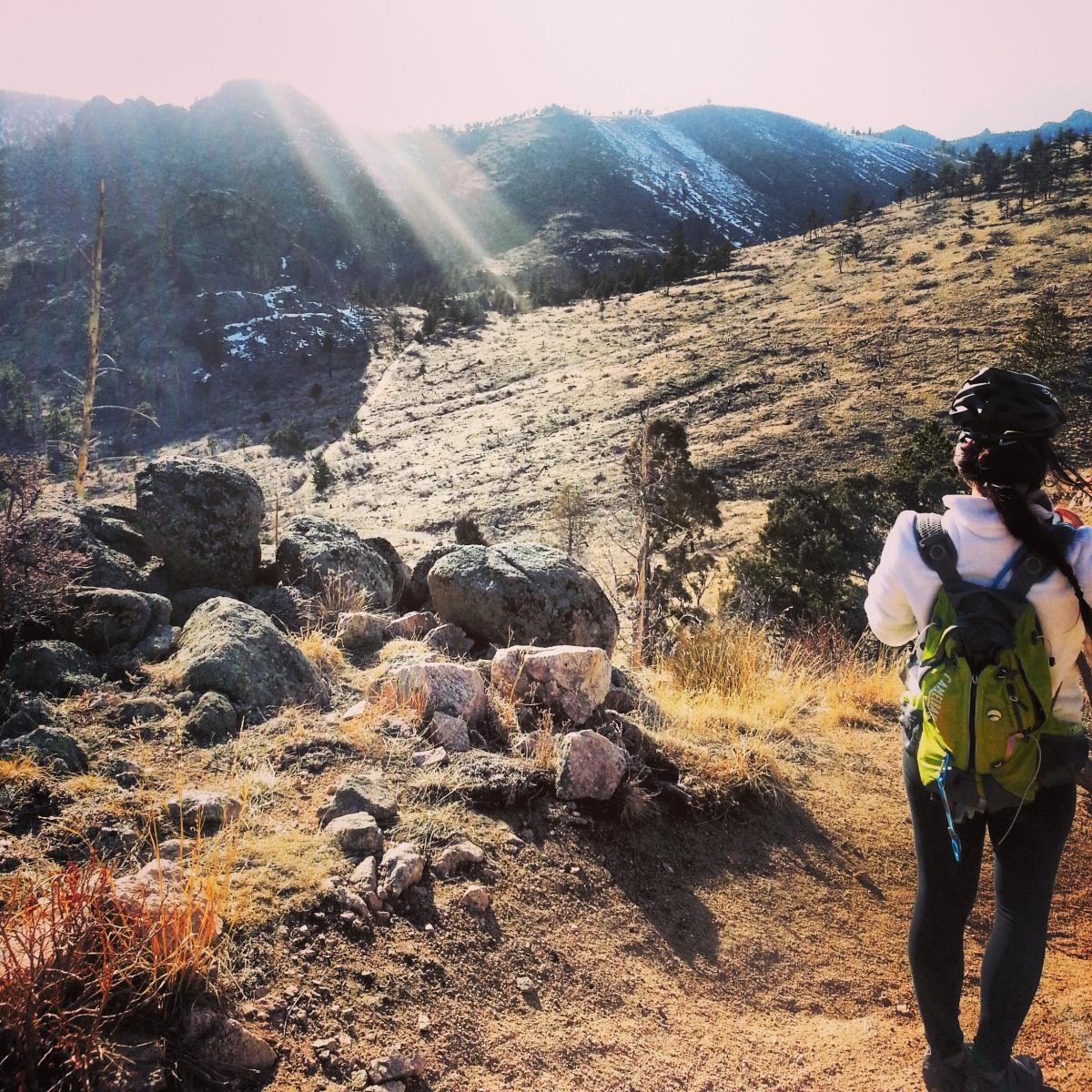 A person wearing a helmet and a backpack stands on a rocky trail, gazing at a mountainous landscape bathed in sunlight. The serene scene features rolling hills with patches of grass and sparse trees, highlighting the natural beauty of the outdoors. Walker Ranch mountain bike trail.