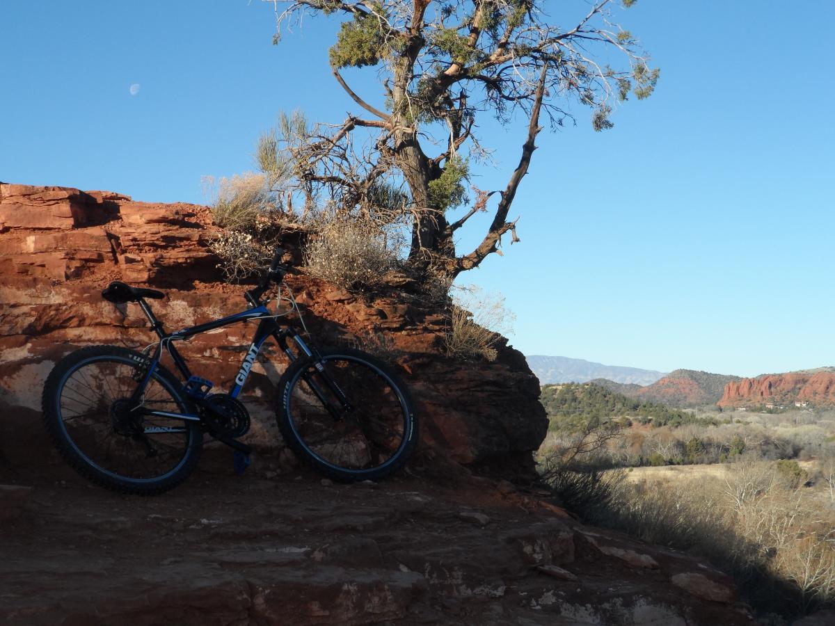 Giant Revel 2: A blue mountain bike leaning against a rocky outcrop, with a clear blue sky above and a crescent moon visible. In the background, there are rolling hills and a landscape of sparse vegetation, showcasing the natural beauty of the area.