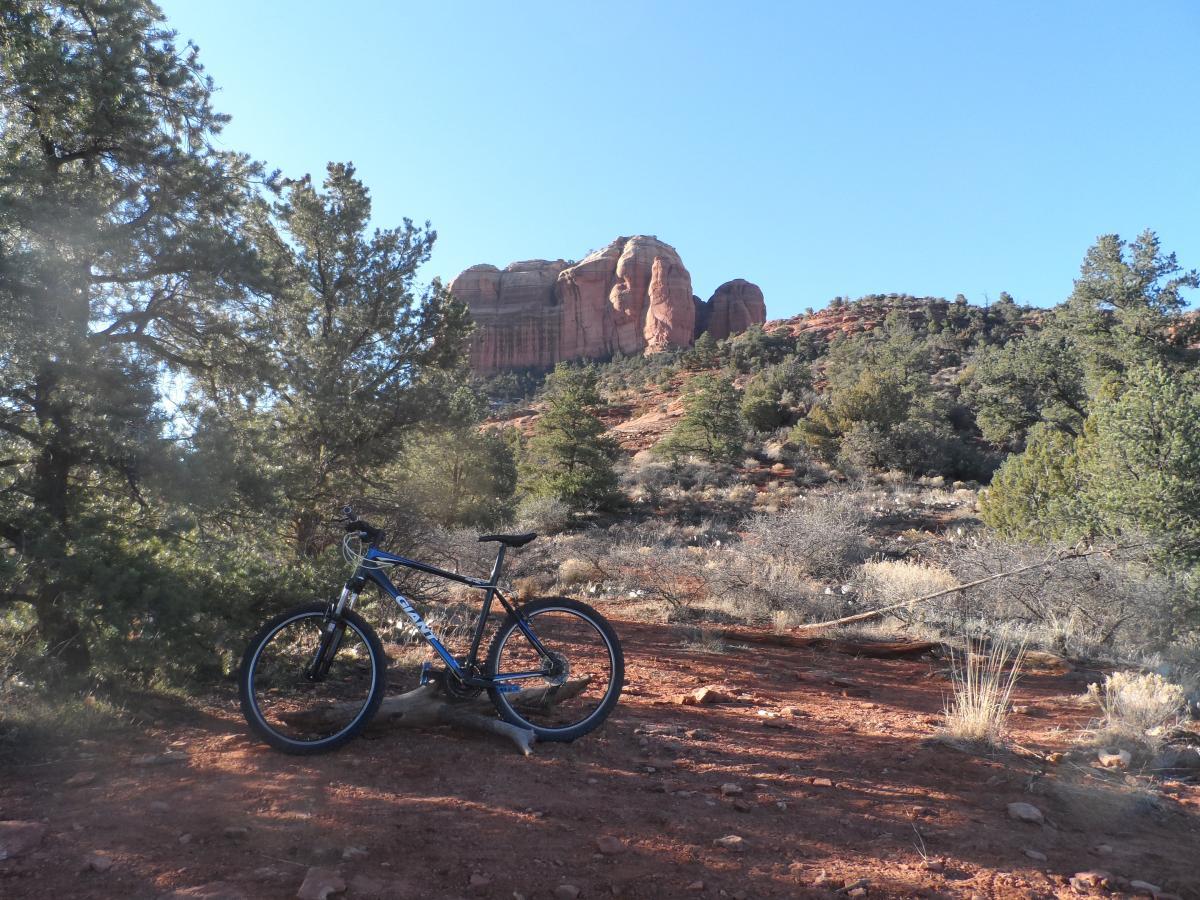 Giant Revel 2: A mountain bike rests on a dirt path surrounded by shrubs and trees, with red rock formations in the background under a clear blue sky.