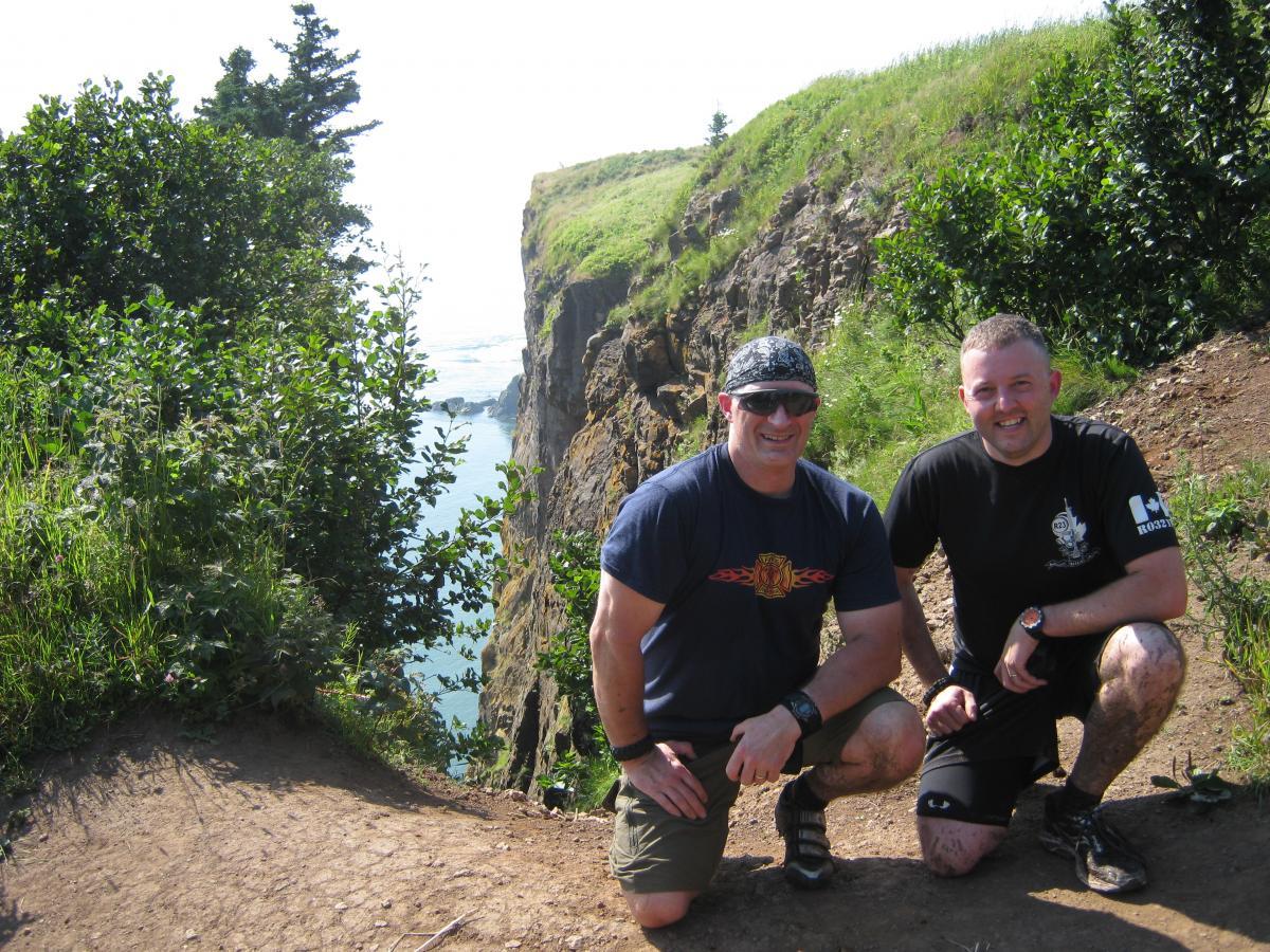 Two men kneel on a dirt path with a view of a cliff and ocean in the background. They are surrounded by greenery and appear to be enjoying a sunny day outdoors. One man is wearing a black t-shirt with a design and khaki shorts, while the other is in a navy blue shirt and also wearing shorts. Both men are smiling and appear to be in a relaxed, casual setting. Cape Split Trail mountain bike trail.