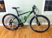 Gary Fisher X-Caliber: A black and green mountain bike leaning against a wall, with a wooden floor and two windows in the background. The bike features thick tires, a lightweight frame, and a suspension system.