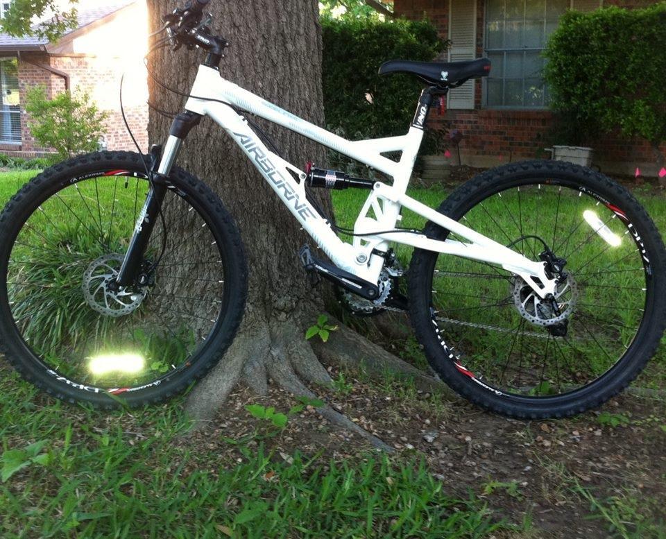Airborne Zeppelin Elite: Mountain bike leaning against a tree, featuring a white frame and black wheels. The bike has disc brakes and reflective elements on the tires, with a grassy area and a brick house in the background.