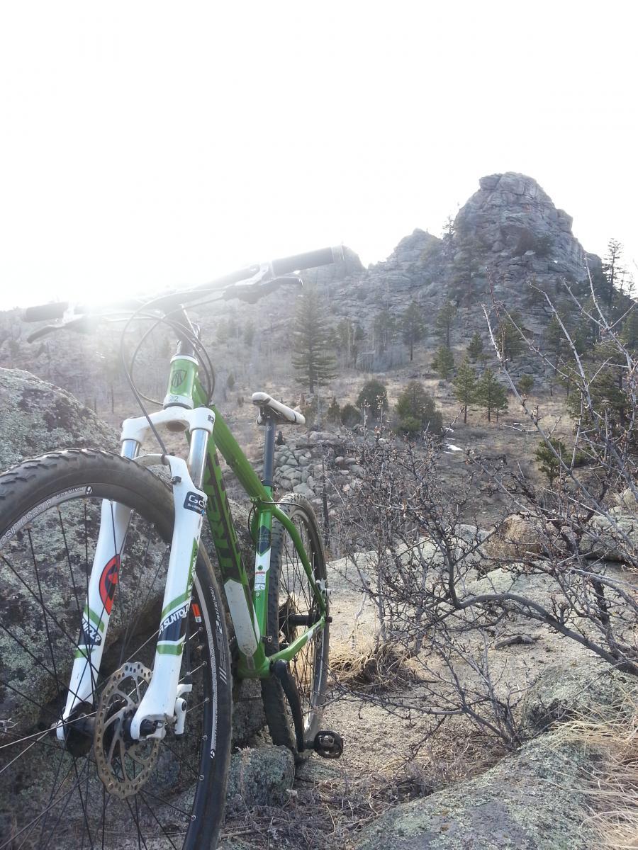 A green mountain bike leaning against a rock, with mountainous terrain and sparse vegetation in the background. The sun is shining, creating a bright atmosphere. Walker Ranch mountain bike trail.