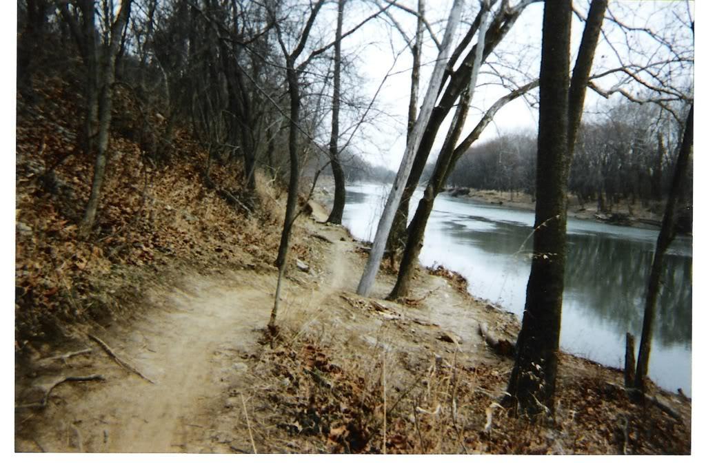 A winding dirt path alongside a calm river, bordered by leafless trees and sparse vegetation. The scene captures a tranquil, slightly overcast day, with the river reflecting the muted colors of the surroundings. Castlewood State Park mountain bike trail.