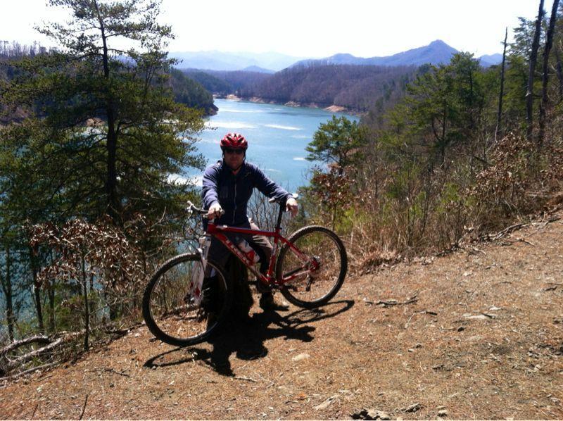 A person wearing a red helmet and a gray jacket stands next to a red mountain bike on a dirt path overlooking a lake surrounded by mountains and trees. The scene is sunny, showcasing a vibrant outdoor landscape. Tsali Right Loop mountain bike trail.