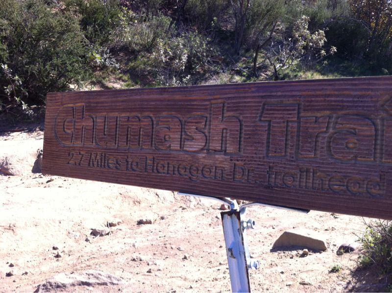 Wooden sign for Chumash Trail, indicating a distance of 2.7 miles to Hangon Dr. trailhead, set against a natural landscape. Chumash mountain bike trail.