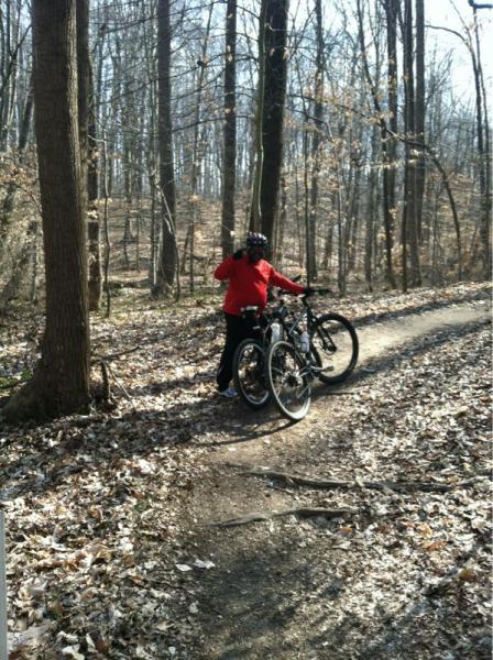 A person in a red jacket stands next to two mountain bikes on a dirt trail in a forested area. The surroundings feature bare trees with a few remaining leaves, suggesting early spring or late autumn. Sunlight filters through the tree branches, casting gentle shadows on the ground. Rosaryville State Park mountain bike trail.
