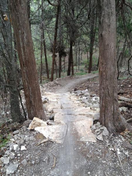 A rocky pathway descending through a wooded area, flanked by tall trees. The trail is partially lined with stone steps and surrounded by scattered rocks, leading into a lush green forest in the background. Big Cedar Wilderness Trails mountain bike trail.