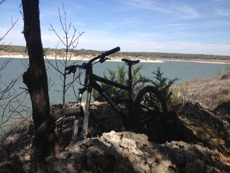 A mountain bike leaning against a rocky outcrop with a scenic view of a lake and green hills in the background under a clear blue sky. Goodwater Trail mountain bike trail.