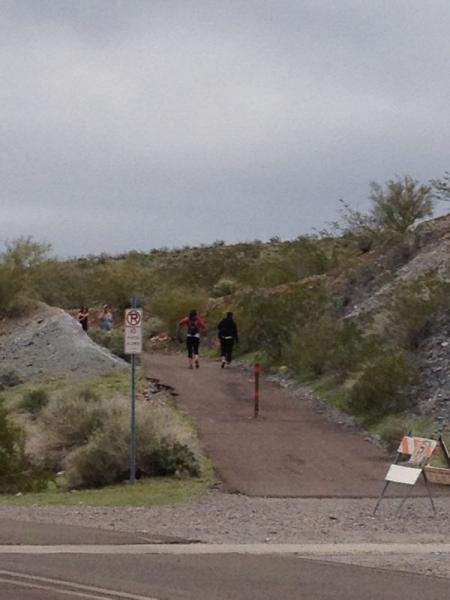 A group of people running along a trail in a desert landscape, surrounded by shrubs and rocky terrain under a cloudy sky. A sign indicating no parking is visible in the foreground. Trail #100 mountain bike trail.