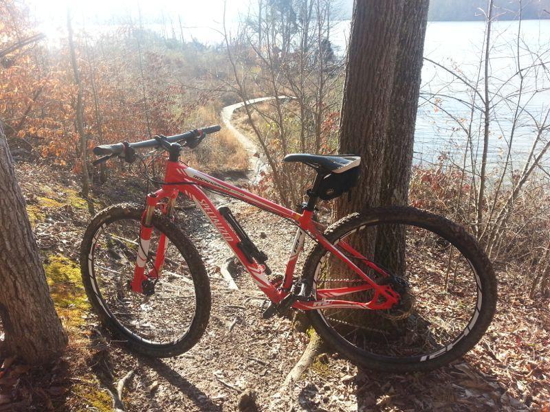 A red mountain bike leaning against a tree on a dirt path that winds along a lake, surrounded by trees and autumn foliage. The sun is shining, creating a bright and inviting atmosphere. Haw Ridge Park mountain bike trail.