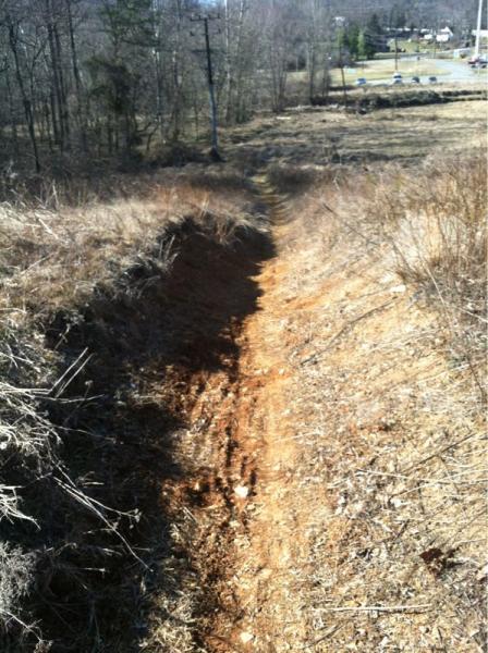 A narrow, dirt path running through a dry, grassy area, bordered by sparse vegetation and trees. The path shows signs of erosion, with an uneven surface and embankments on either side. In the background, there are signs of a parking lot and other structures among the trees. Wakefield mountain bike trail.