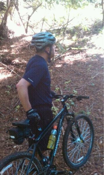 A person wearing a bicycle helmet and gloves stands next to a mountain bike on a trail surrounded by trees. The individual is looking away, with one hand resting on the bike's seat. A water bottle is attached to the bike. The ground is covered in leaves and the setting appears to be sunny and forested. Wilder Ranch State Park mountain bike trail.