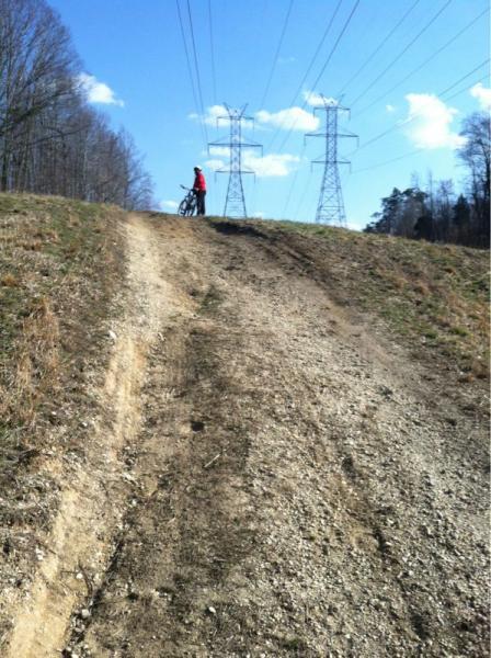 A cyclist stands at the top of a steep, gravelly hill with power lines in the background under a clear blue sky. The path is uneven and worn, suggesting frequent use for biking or hiking. Rosaryville State Park mountain bike trail.