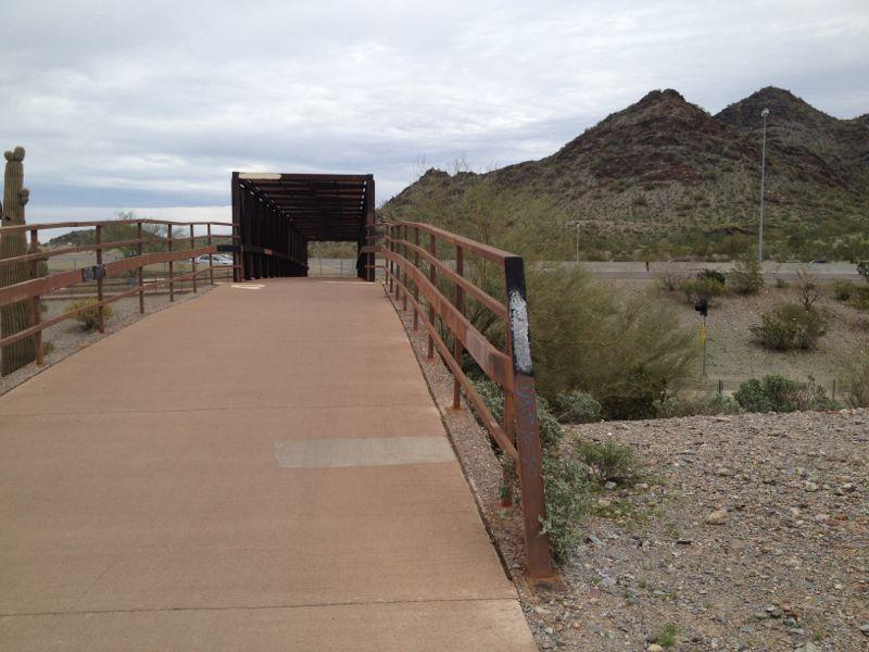 A paved walkway with metal railings leads towards a covered structure, surrounded by desert vegetation and rolling hills in the background. The sky is overcast, and a road is visible in the distance. Trail #100 mountain bike trail.