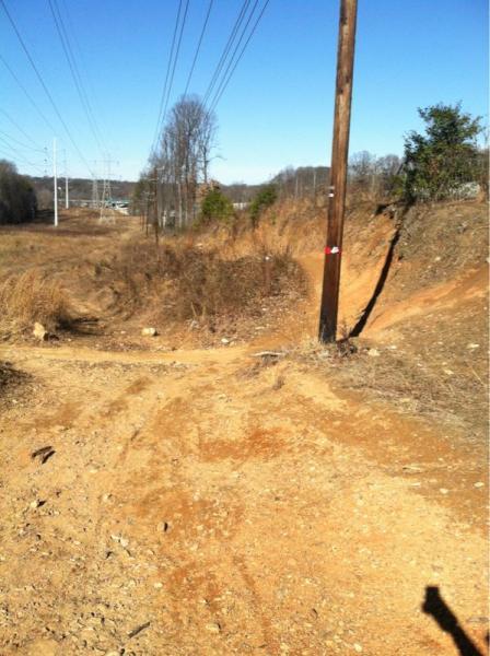 A dirt path winding through an open area, flanked by dry vegetation and a wooden utility pole. Power lines stretch across the sky in the background. The landscape is mostly bare with a few scattered rocks and shrubs, under a clear blue sky. Wakefield mountain bike trail.