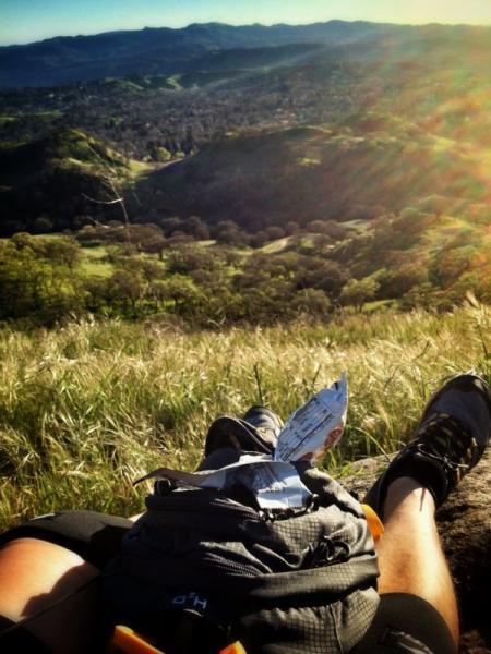 A person resting on a rocky outcrop with their legs extended, wearing hiking shoes. In the foreground, a backpack is positioned near their feet. The background features a sweeping view of green hills and valleys under a bright blue sky, illuminated by sunlight, creating a serene outdoor atmosphere. Shell Ridge Open Space mountain bike trail.