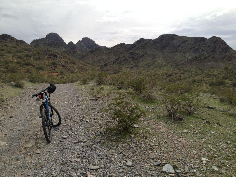 A mountain bike rests on a rocky trail surrounded by green vegetation and rugged hills under a cloudy sky. Trail #100 mountain bike trail.