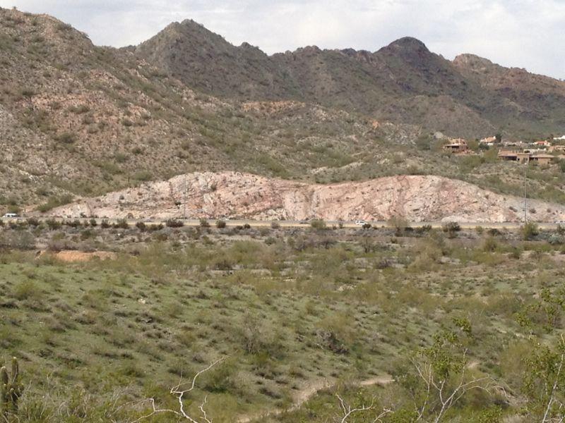 A scenic view of rocky terrain and rolling hills under a partly cloudy sky, with sparse vegetation and a road running through the foreground. The landscape features a mix of light-colored rock formations and green shrubbery, and distant houses are visible on the hillside. Trail #100 mountain bike trail.