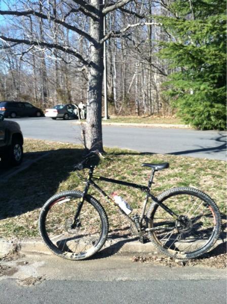 A black mountain bike with a visible layer of dirt parked beside a tree, with a gravel area and a road in the background. A few cars can be seen parked in the distance, and the scene is set in a wooded area with bare trees. Wakefield mountain bike trail.