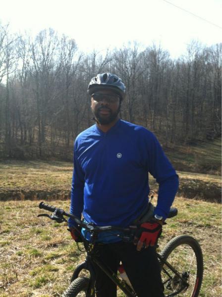 A person wearing a blue long-sleeve shirt and a black helmet stands beside a mountain bike. They are outdoors in a grassy area with trees in the background. The weather appears sunny, suggesting a pleasant day for biking. Rosaryville State Park mountain bike trail.