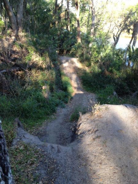 A dirt path winding through a wooded area, featuring greenery and trees on either side. The trail slopes downward, leading toward a body of water visible in the background. Sunlight filters through the foliage, creating dappled shadows on the ground. Grapefruit Trail mountain bike trail.