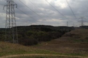 A landscape featuring power lines running across a grassy area with a path, set against a backdrop of hills and a cloudy sky. Big Cedar Wilderness Trails mountain bike trail.
