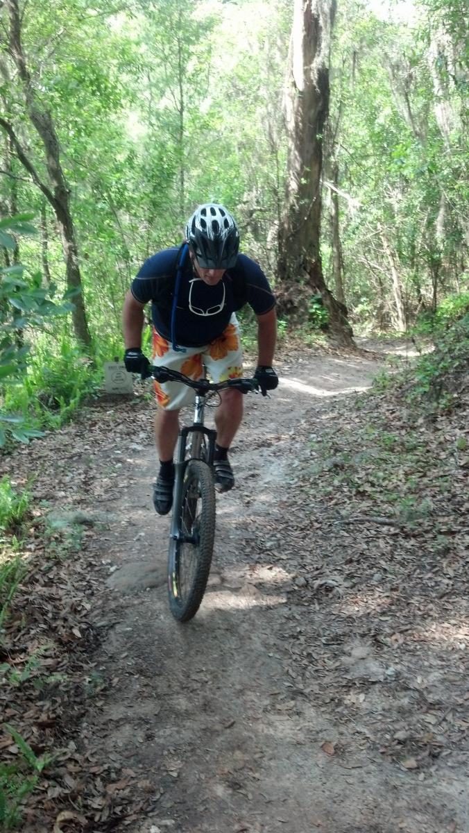 A person in a helmet and cycling gear rides a mountain bike along a dirt trail surrounded by lush greenery. The cyclist is leaning forward, navigating the winding path through a forested area. Balm Boyette Scrub Preserve mountain bike trail.