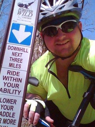 A person in a bright yellow cycling jersey smiles while posing next to a trail sign that indicates a downhill section for the next three miles, advising cyclists to ride within their ability. The background shows a wooded area, and the cyclist is wearing sunglasses and a helmet. Coldwater Mountain mountain bike trail.