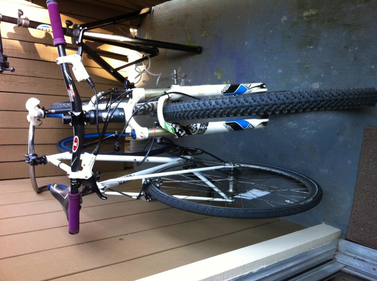 Trek X-Caliber: A close-up image of a bicycle leaning against a wall, primarily showing the handlebars, front fork, and front wheel. The bike has a white frame with some black and blue detailing, and the handlebars feature a purple grip. The image captures the bike on a wooden floor near a doorway.