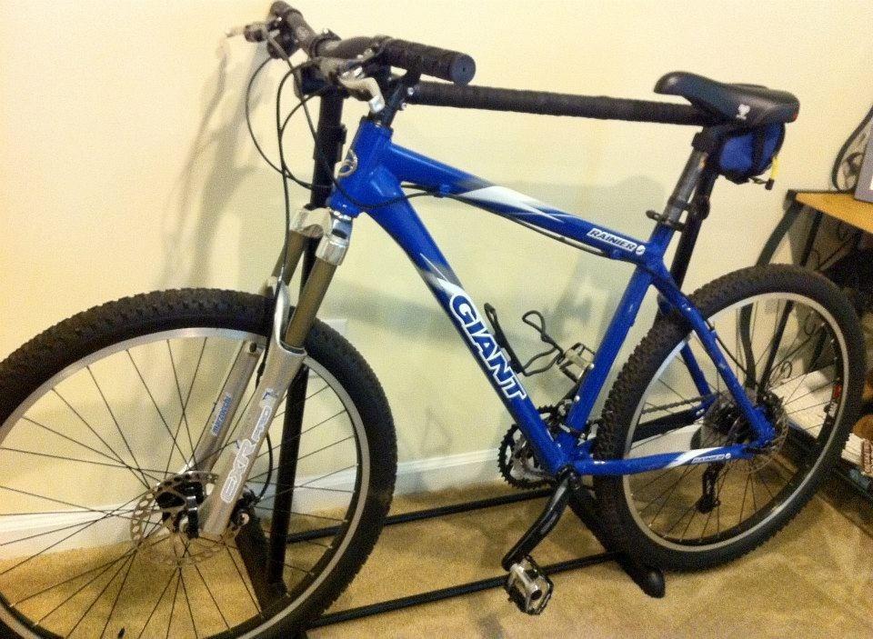 Giant Ranier: A blue mountain bike leaning on a bike stand, featuring thick tires, a front suspension fork, and a black handlebar with grips. The bike is partially in focus, with a plain wall in the background and a small table visible nearby.