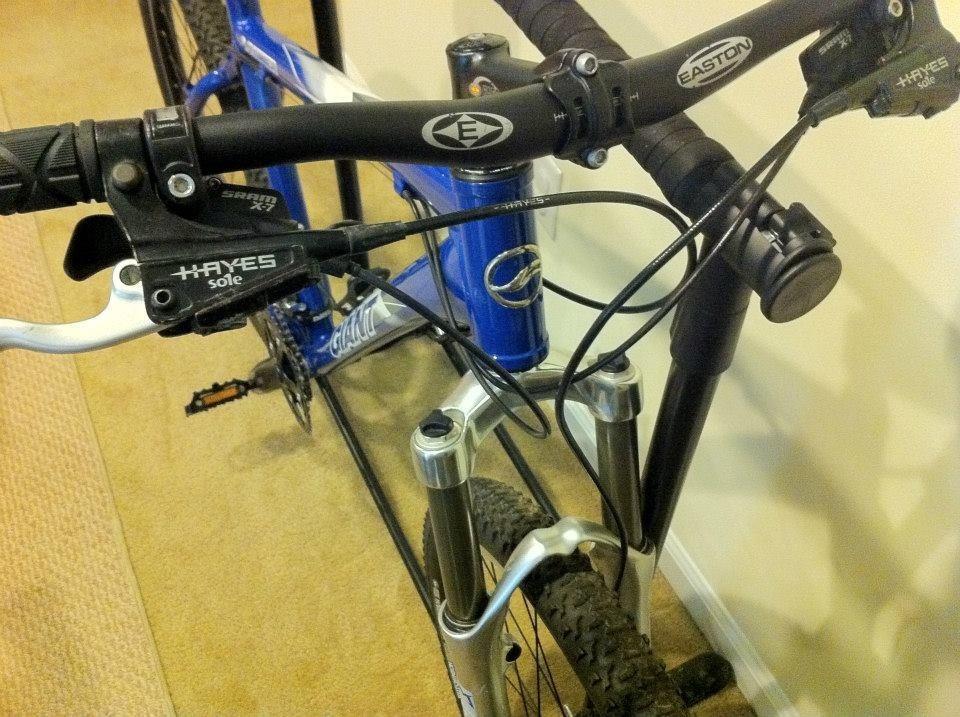 Giant Ranier: Close-up view of a mountain bike's handlebars and front suspension, featuring branded components including brake levers and cables. The bike has a blue frame with a visible logo and is positioned in a home environment, highlighting the textured surface of the floor.