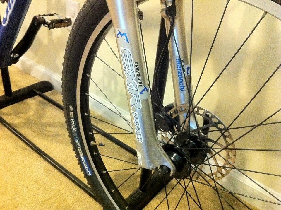Giant Ranier: Close-up of a bicycle front wheel, showcasing the alloy fork labeled "Marzocchi EXR Pro," a disc brake, and spokes. The wheel is positioned on a floor stand against a neutral-colored wall and carpet.