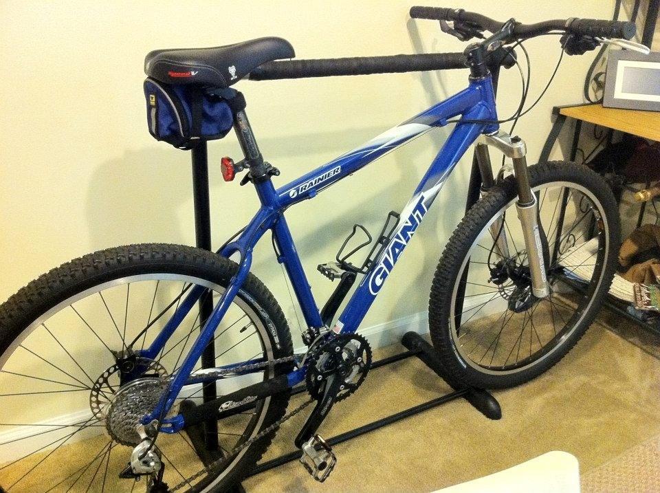 Giant Ranier: A blue Giant mountain bike displayed on a bike stand inside a room, with a black seat and a small bag attached. The ground is carpeted, and there are various items visible in the background, including a microwave on a table.