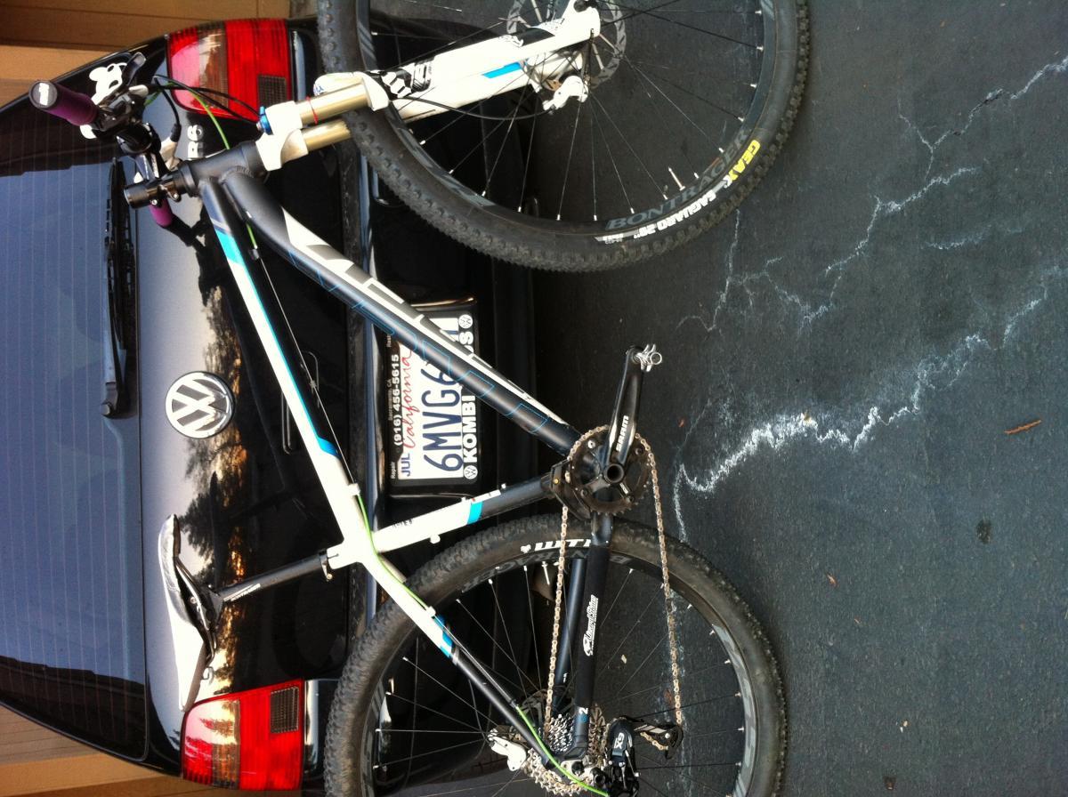 Trek X-Caliber: A mountain bike leaning against a black Volkswagen car, with the side view showing the bike’s frame, handlebar, and rear wheel. The car's license plate is partially visible, along with the reflection of clouds in the vehicle's surface. The ground surface shows light markings, and the scene is set in an outdoor parking area.