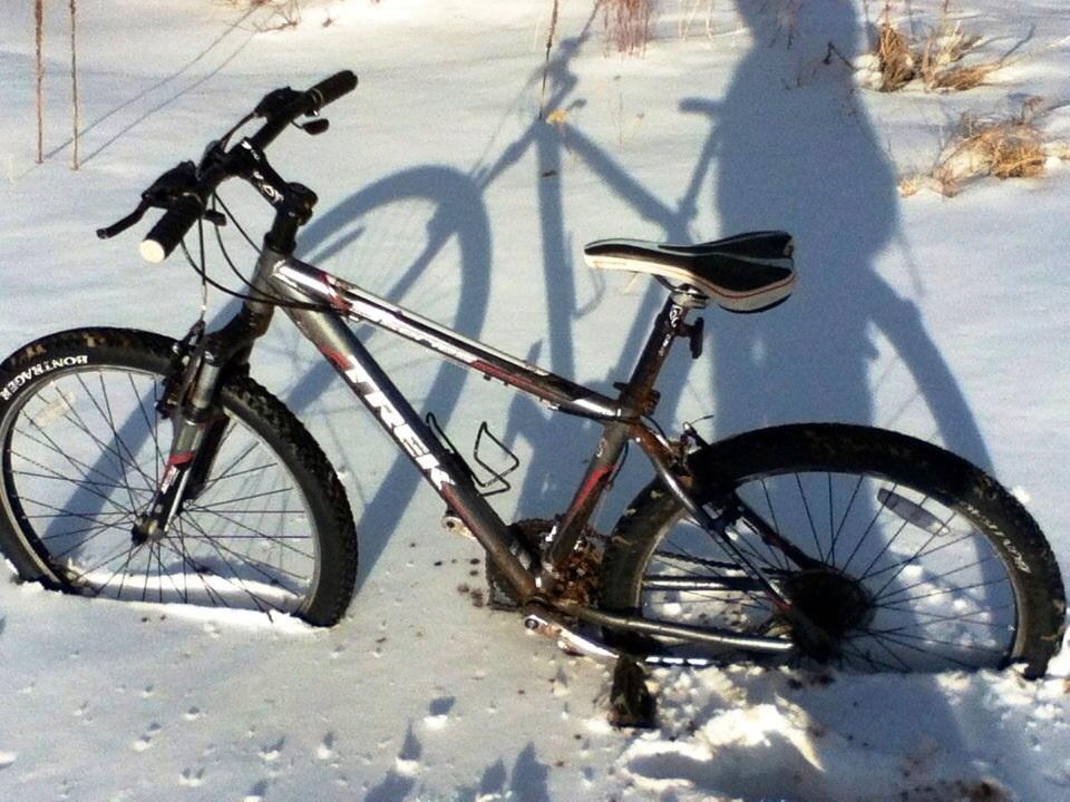 Trek 4500: A mountain bike parked on a snowy ground, with its shadow visible in the sunlight. The bike has a silver frame, black handlebars, and a white seat, with muddy tires showing signs of use. Snow-covered terrain surrounds the bike, indicating winter conditions.