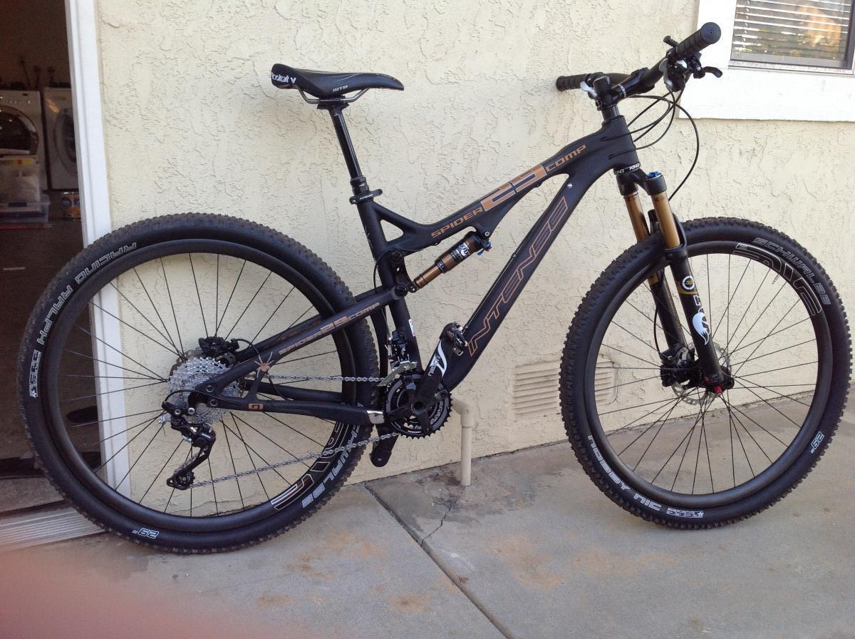Intense Spider 29 Comp: A black mountain bike with dual suspension, featuring thick tires and a modern frame design, stands against a textured beige wall. The bike is equipped with a Shimano drivetrain, disc brakes, and a sleek saddle. Sunlight reflects off the bike's components, highlighting its features. In the background, there are hints of a laundry area and a window with blinds.