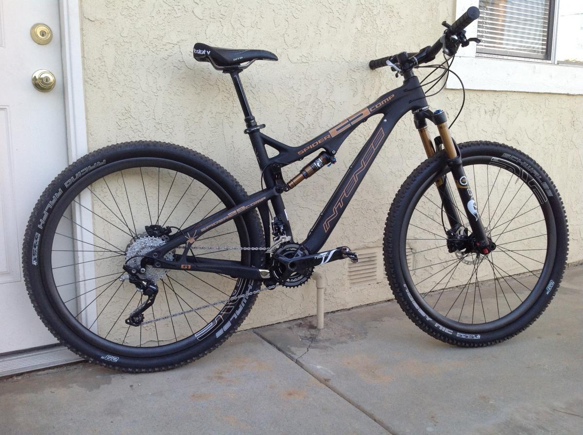 Intense Spider 29 Comp: A black mountain bike with gold accents and thick tires, parked next to a door with a textured wall in the background. The bike features a suspension fork and a gear system visible near the pedals.
