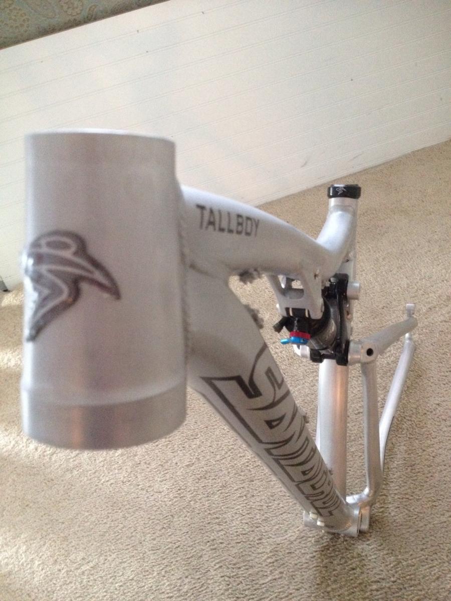 Santa Cruz Tallboy: Alt text: Close-up of a silver bicycle frame with the text "TALLBOY" visible on the downtube, resting on a carpeted surface. The frame has a distinctive design and logo on the head tube.