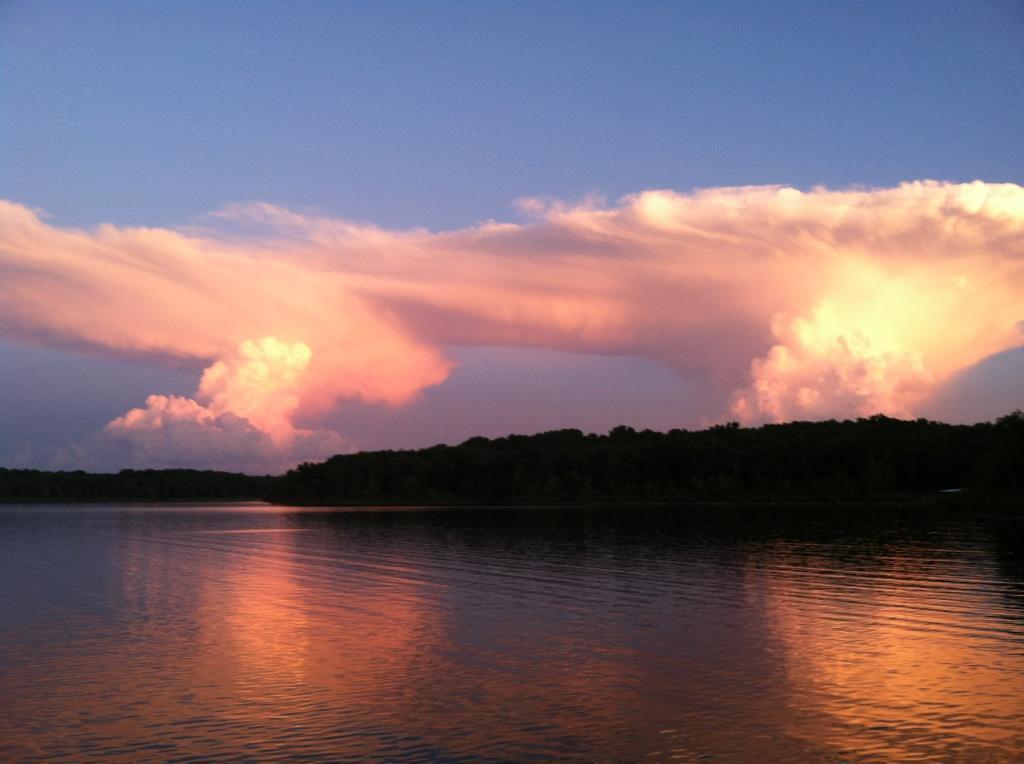 A serene view of a lake at sunset, featuring clouds in shades of pink and orange reflecting beautifully on the water's surface. The horizon is lined with trees, creating a peaceful natural landscape. Rudledge Bluff Overlook mountain bike trail.