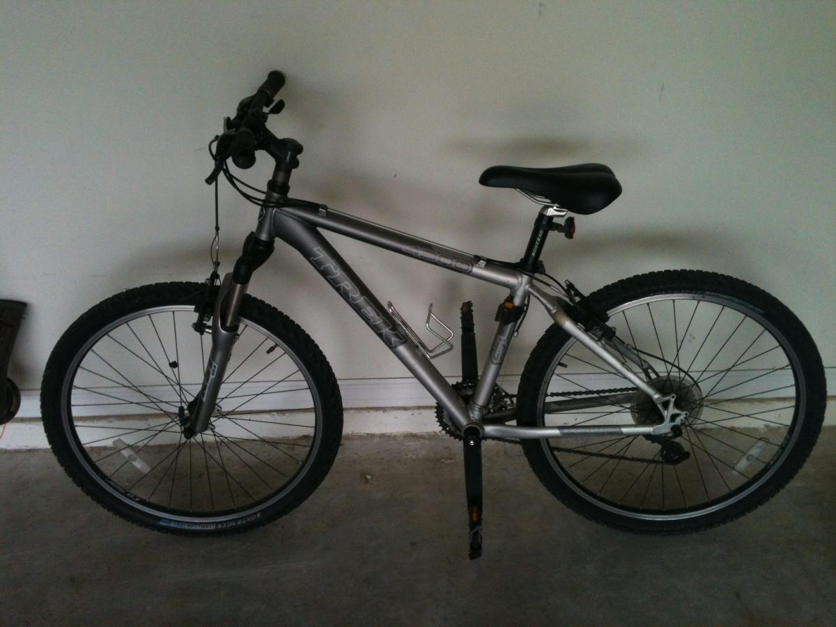 Trek 4300: A silver mountain bike leaning against a plain wall, featuring two wheels with thick, knobby tires, handlebars with grips, and a black seat. The bike has visible gears and a water bottle holder, resting on a concrete floor.
