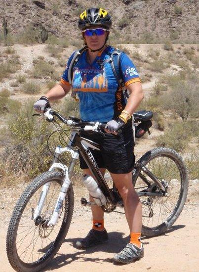 Trek Fuel EX 8: A person standing next to a mountain bike on a dusty trail, wearing a colorful cycling jersey and sunglasses. The background features a desert landscape with sparse vegetation and rocky terrain.